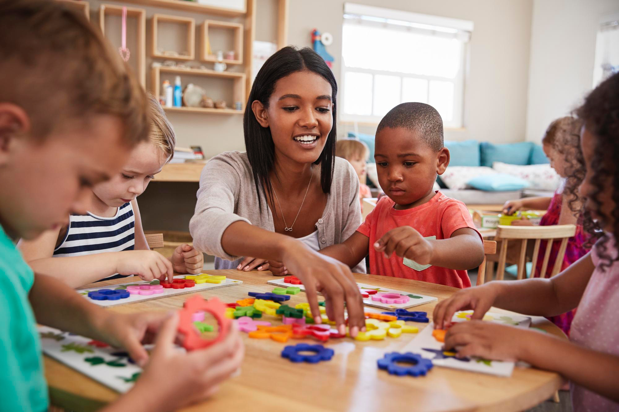 Mujeres sentadas en una pequeña mesa en un aula preescolar con 4 niños pequeños, todos observándola mientras les muestra algo con los bloques de colores sobre la mesa.