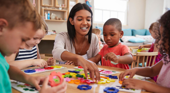 Mujeres sentadas en una pequeña mesa en un aula preescolar con 4 niños pequeños, todos observándola mientras les muestra algo con los bloques de colores sobre la mesa.