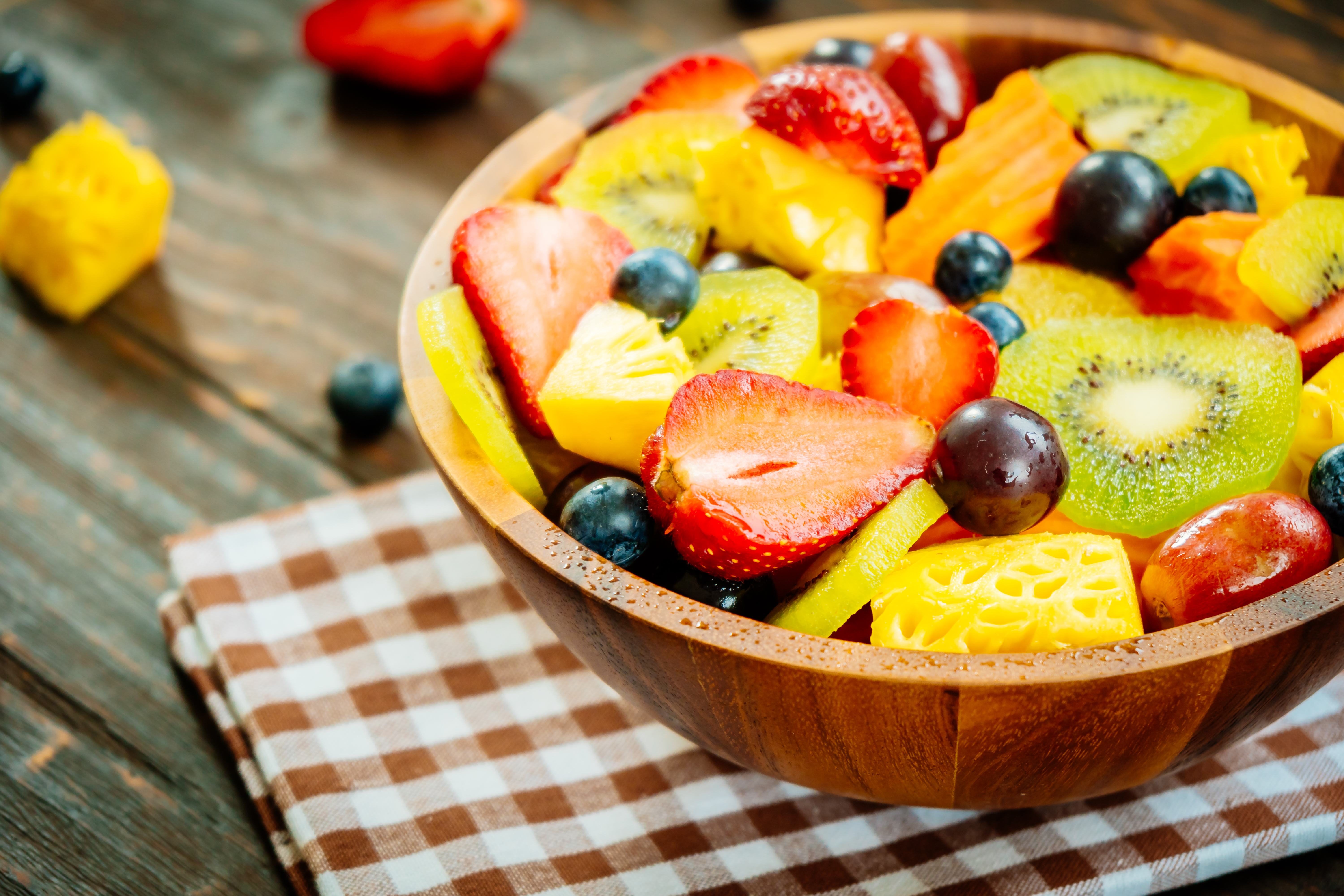 Un plato de frutas variadas que incluyen fresas, arándanos, piña y kiwi. El cuenco está sobre una mesa de madera con un mantel a cuadros debajo.