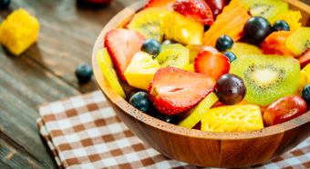 Un plato de frutas variadas que incluyen fresas, arándanos, piña y kiwi. El cuenco está sobre una mesa de madera con un mantel a cuadros debajo.