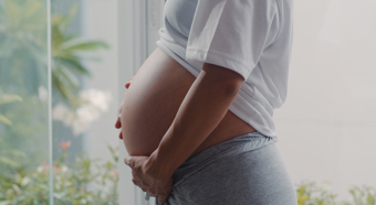 Primer plano de una mujer embarazada sosteniendo suavemente su vientre, vestida con una camiseta blanca y pantalones deportivos grises.