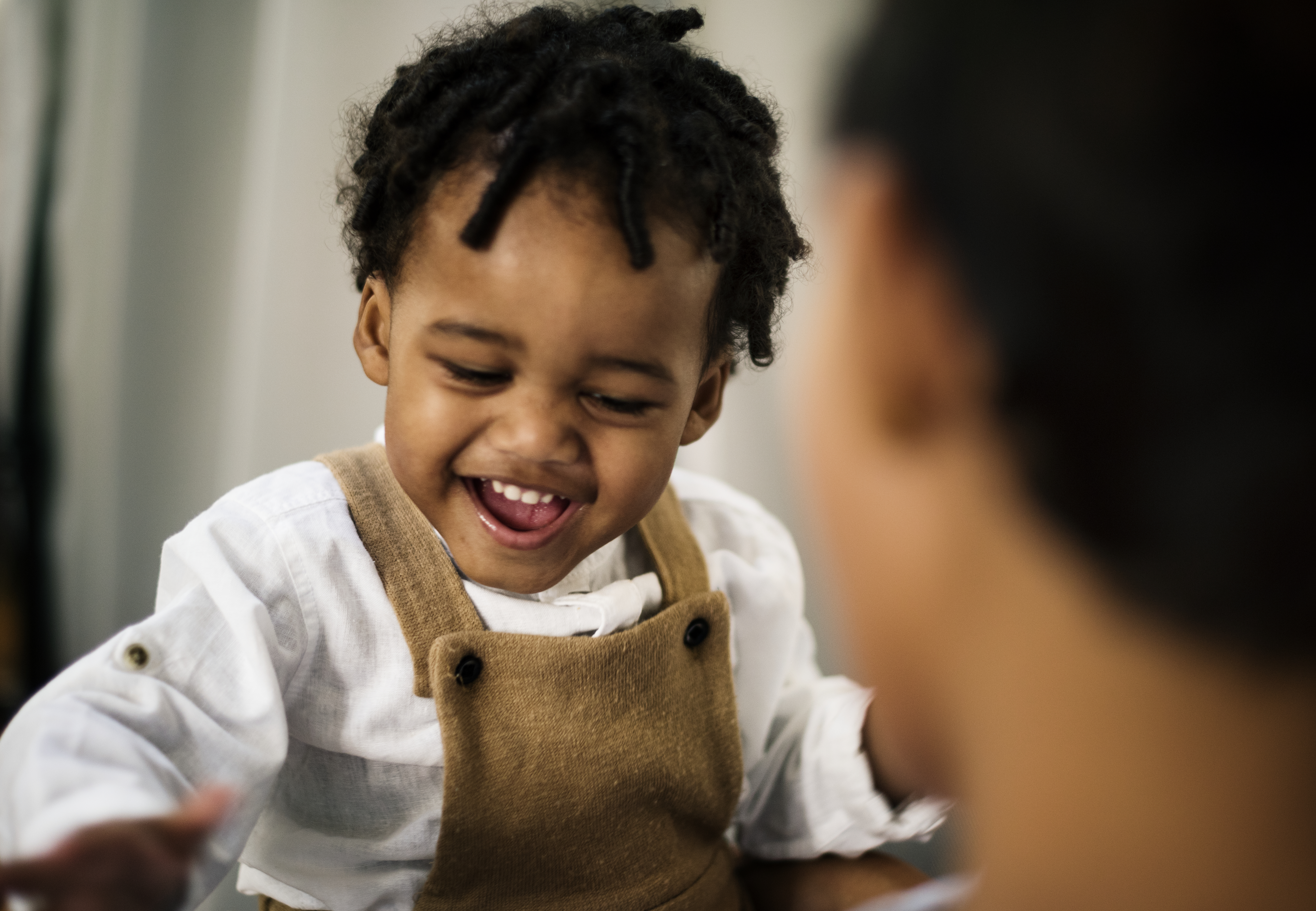 Primer plano de un niño afroamericano riendo y sonriendo, vistiendo un mono marrón y una camisa blanca.