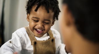 Primer plano de un niño afroamericano riendo y sonriendo, vistiendo un mono marrón y una camisa blanca.