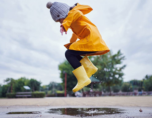 Una niña pequeña vestida con un gorro abrigado, y un piloto y botas de lluvia amarillas, saltando sobre un charco al aire libre, en un día nublado.