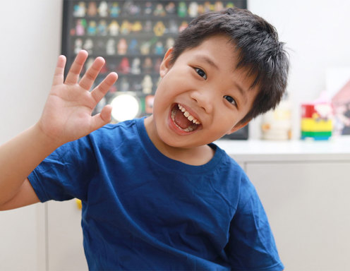 Un niño pequeño de tez marrón claro y pelo oscuro, con una remera azul, sonriéndo y saludando a la cámara con su brazo.