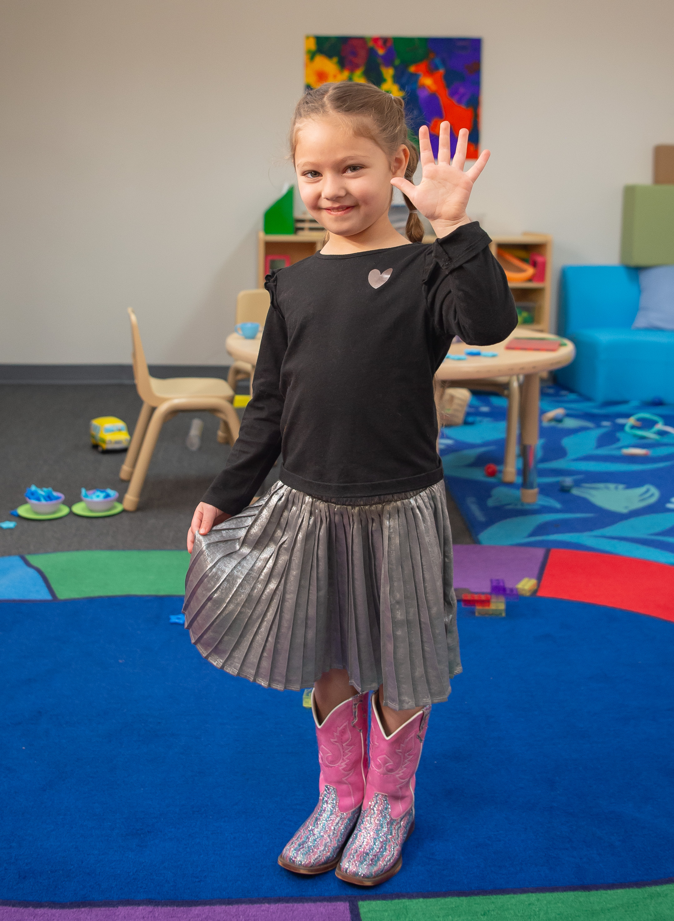 Niña en un entorno de aula colorido, con botas de vaquero rosas y levantando la mano con cinco dedos, sonriendo a la cámara.
