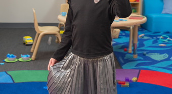 Niña en un entorno de aula colorido, con botas de vaquero rosas y levantando la mano con cinco dedos, sonriendo a la cámara.
