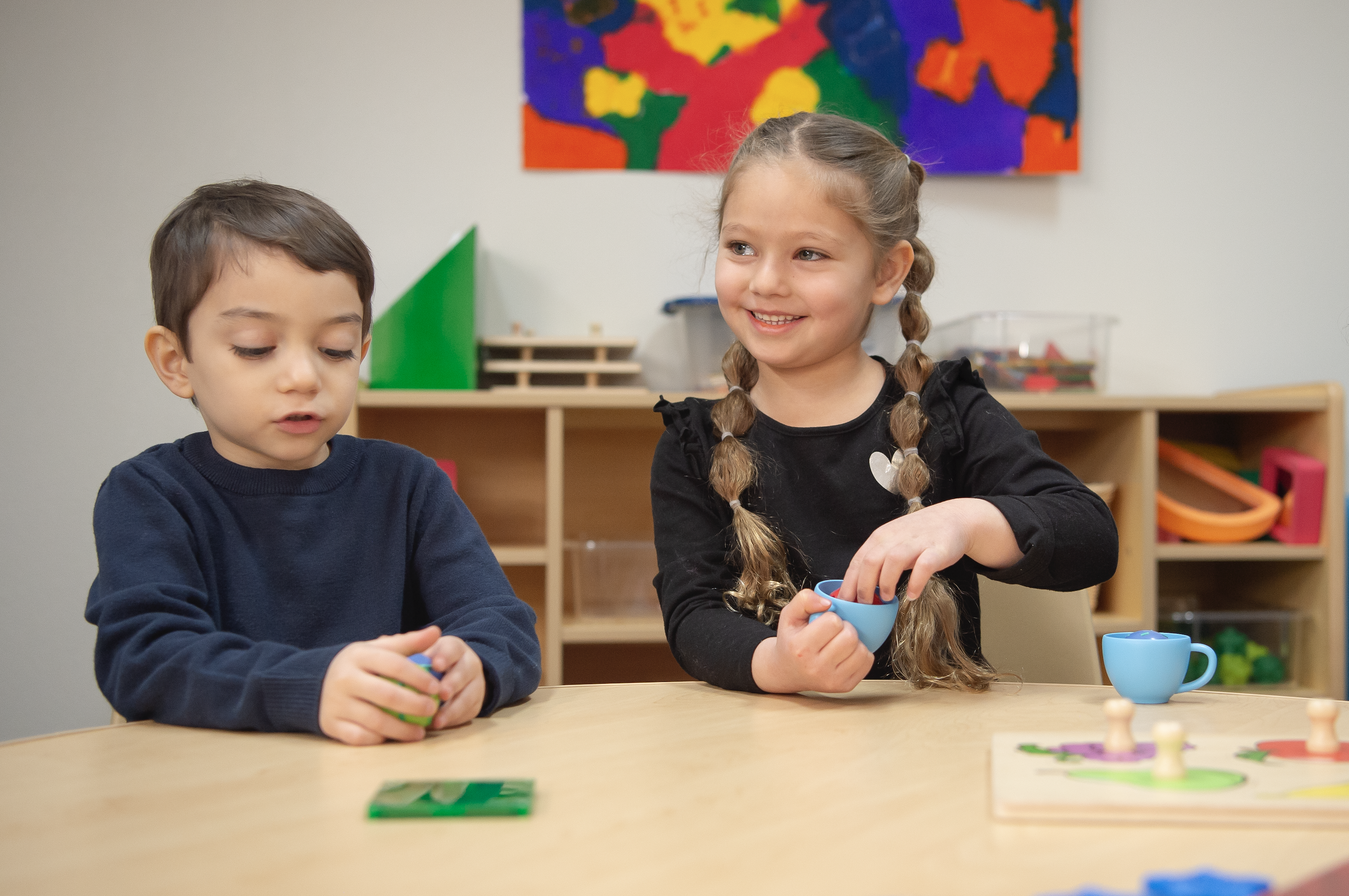 Dos niños pequeños sentados en una mesa en un aula, sonriendo y jugando con juguetes sobre la mesa.