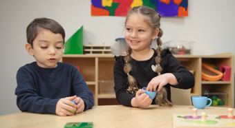 Dos niños pequeños sentados en una mesa en un aula, sonriendo y jugando con juguetes sobre la mesa.