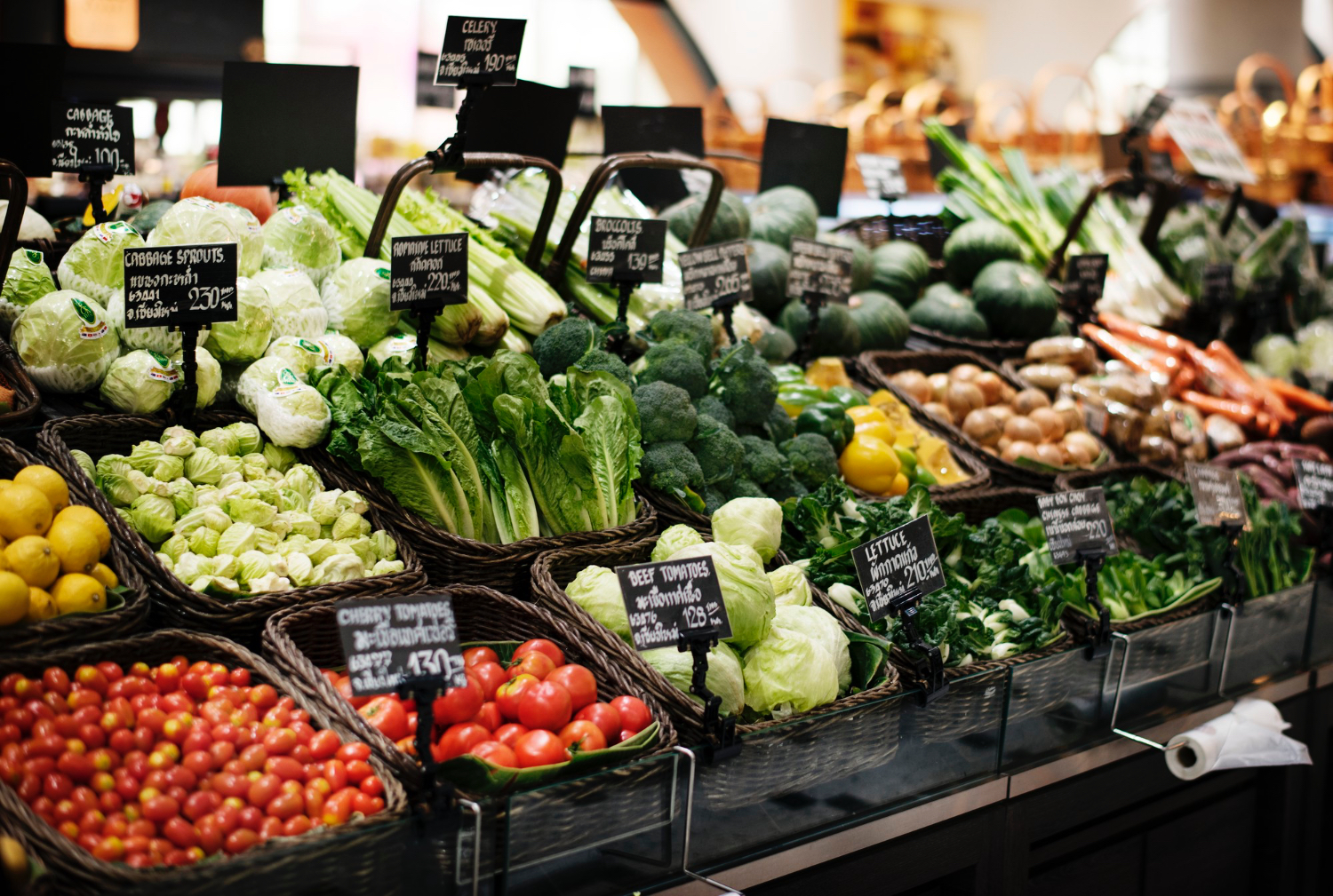 Pasillo de frutas y verduras de un supermercado, con diversos vegetales exhibidos en contenedores. Tomates, lechuga, repollo, zanahorias y más.