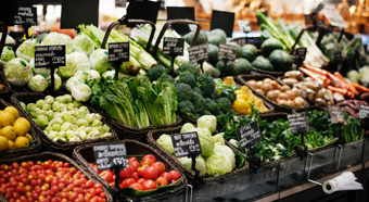 Pasillo de frutas y verduras de un supermercado, con diversos vegetales exhibidos en contenedores. Tomates, lechuga, repollo, zanahorias y más.