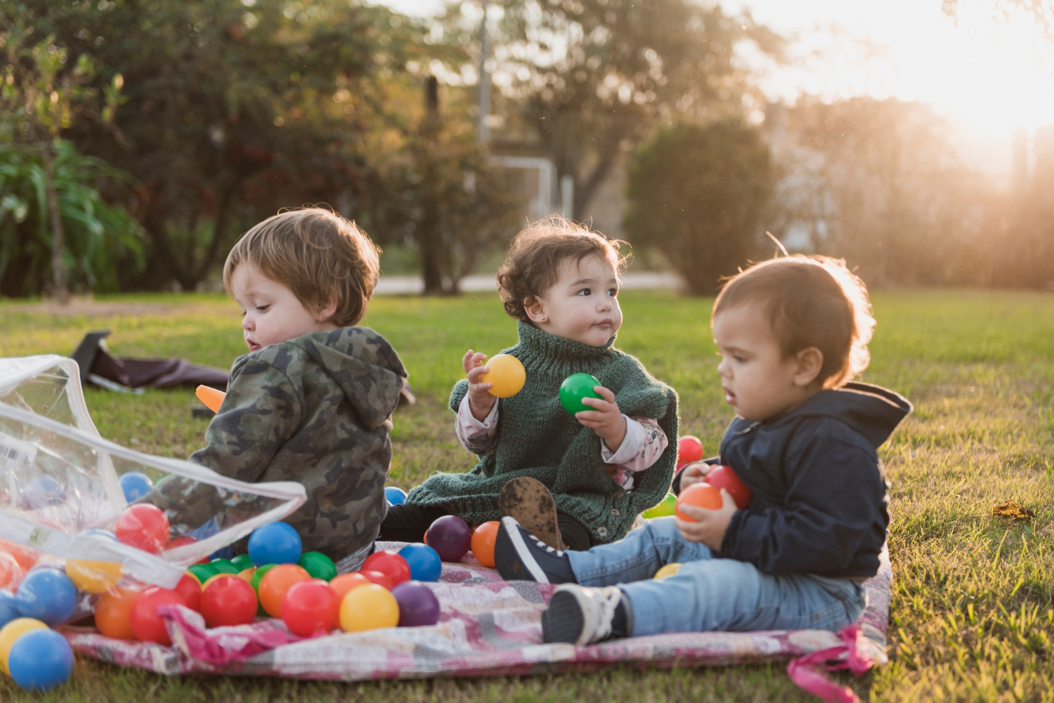 Tres niños pequeños sentados afuera en el césped, jugando con juguetes de colores del arco iris.