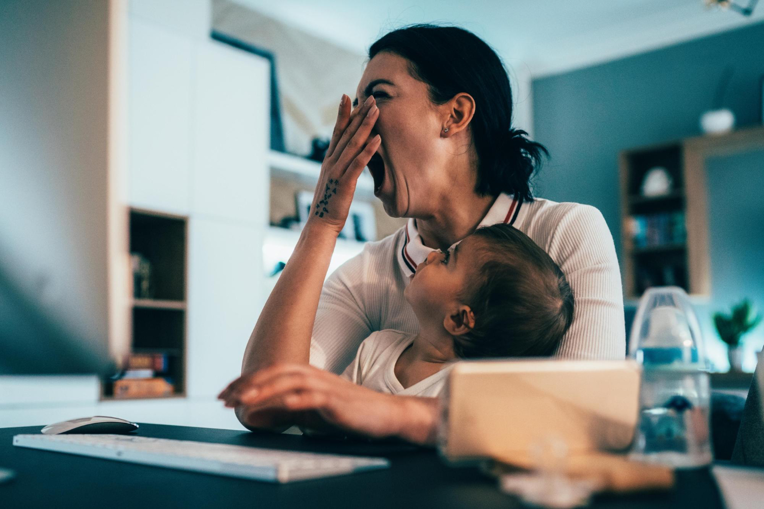 Mujer adulta bostezando, con un niño pequeño en su regazo, sentada en un escritorio en un entorno doméstico.
