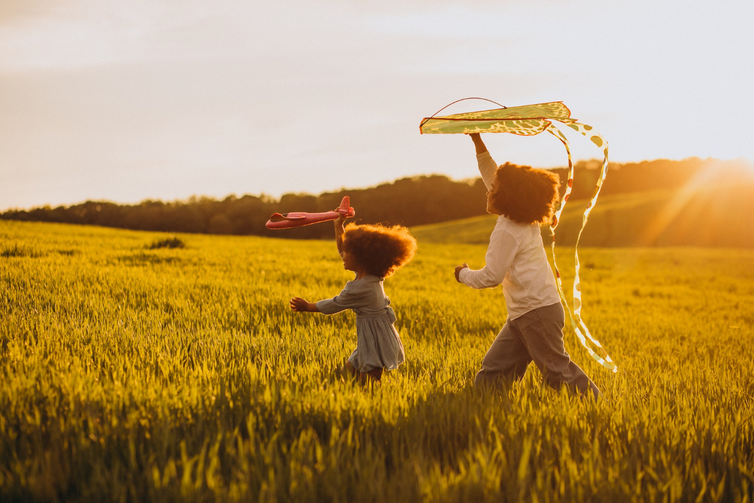 Dos niños pequeños jugando con cometas, mientras corren por un campo de hierba durante la puesta del sol.
