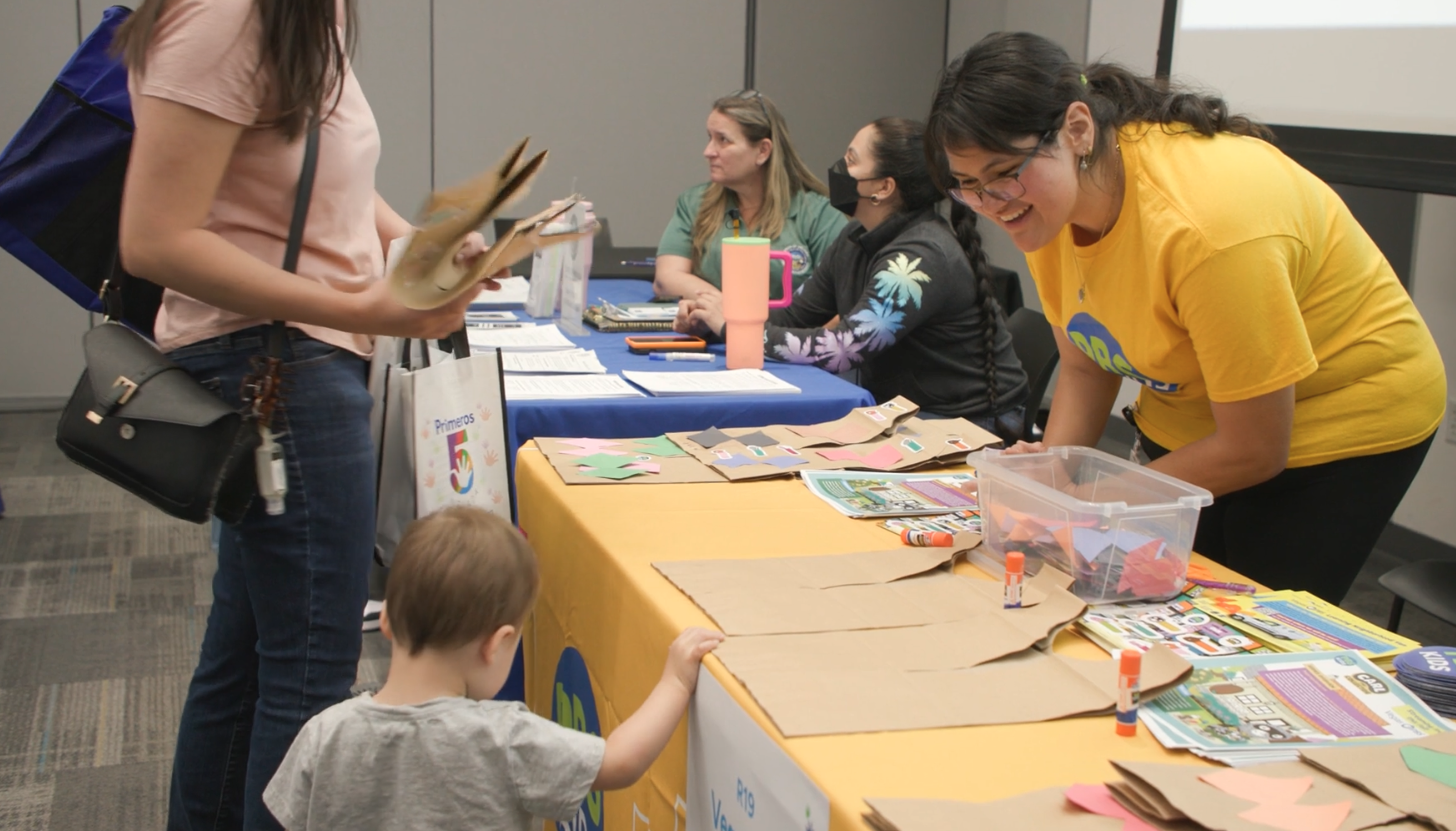 Una mujer que viste una camisa de color amarillo brillante, que trabaja en una mesa en un evento comunitario, sonríe a un niño pequeño y a una mujer adulta que se detienen en su mesa.