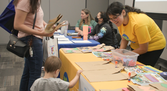 Una mujer que viste una camisa de color amarillo brillante, que trabaja en una mesa en un evento comunitario, sonríe a un niño pequeño y a una mujer adulta que se detienen en su mesa.