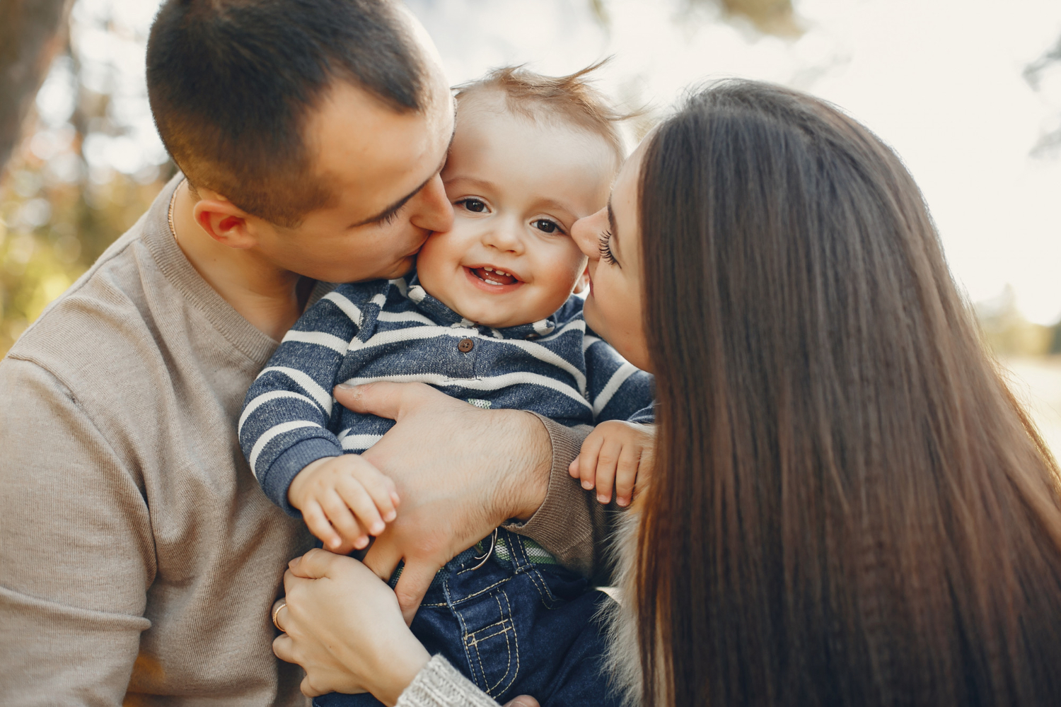 Un niño pequeño sostenido entre dos adultos que besan ambas mejillas del niño sonriente.