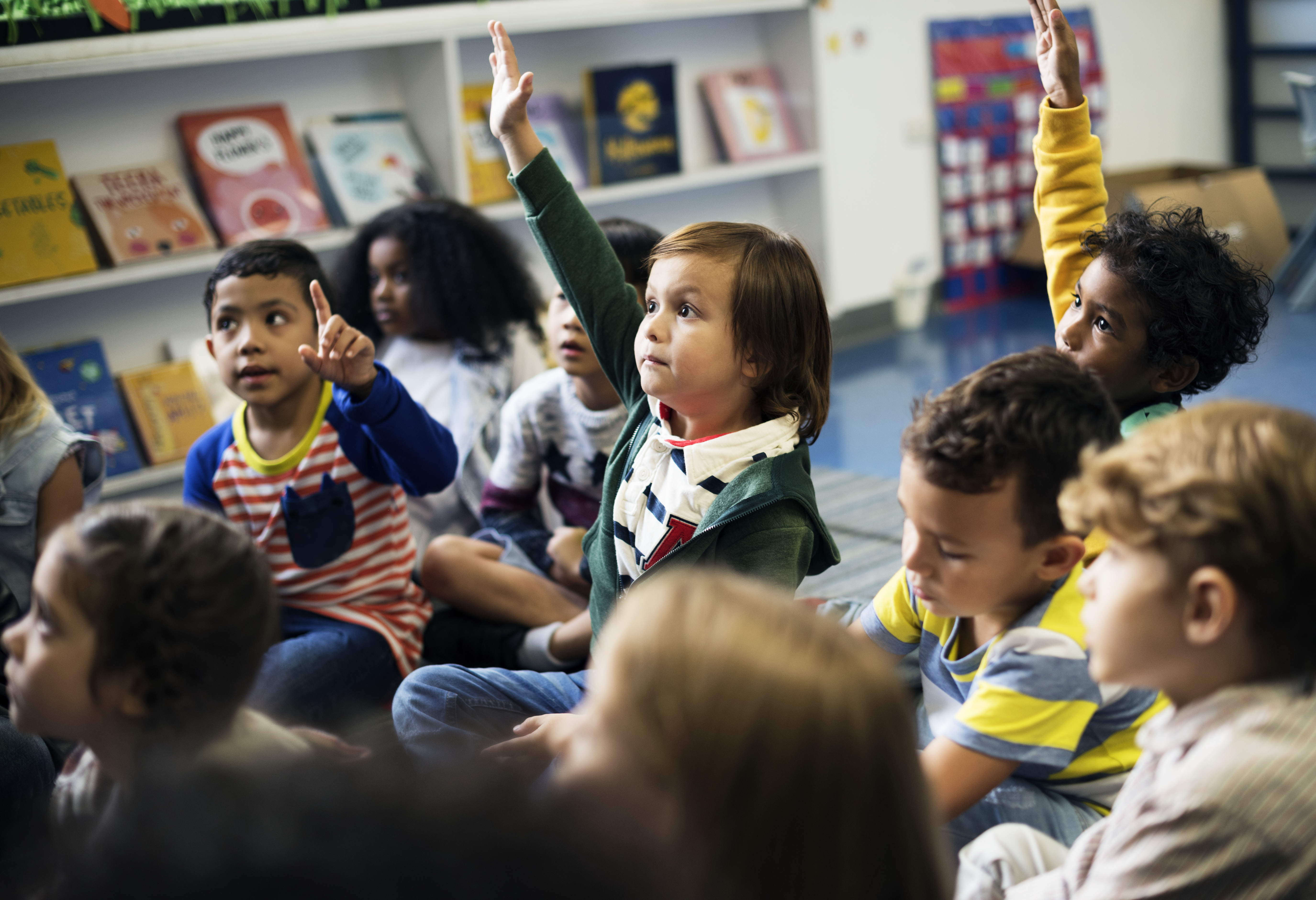 Grupo de niños en un aula sentados en el piso, algunos de ellos levantan sus manos.