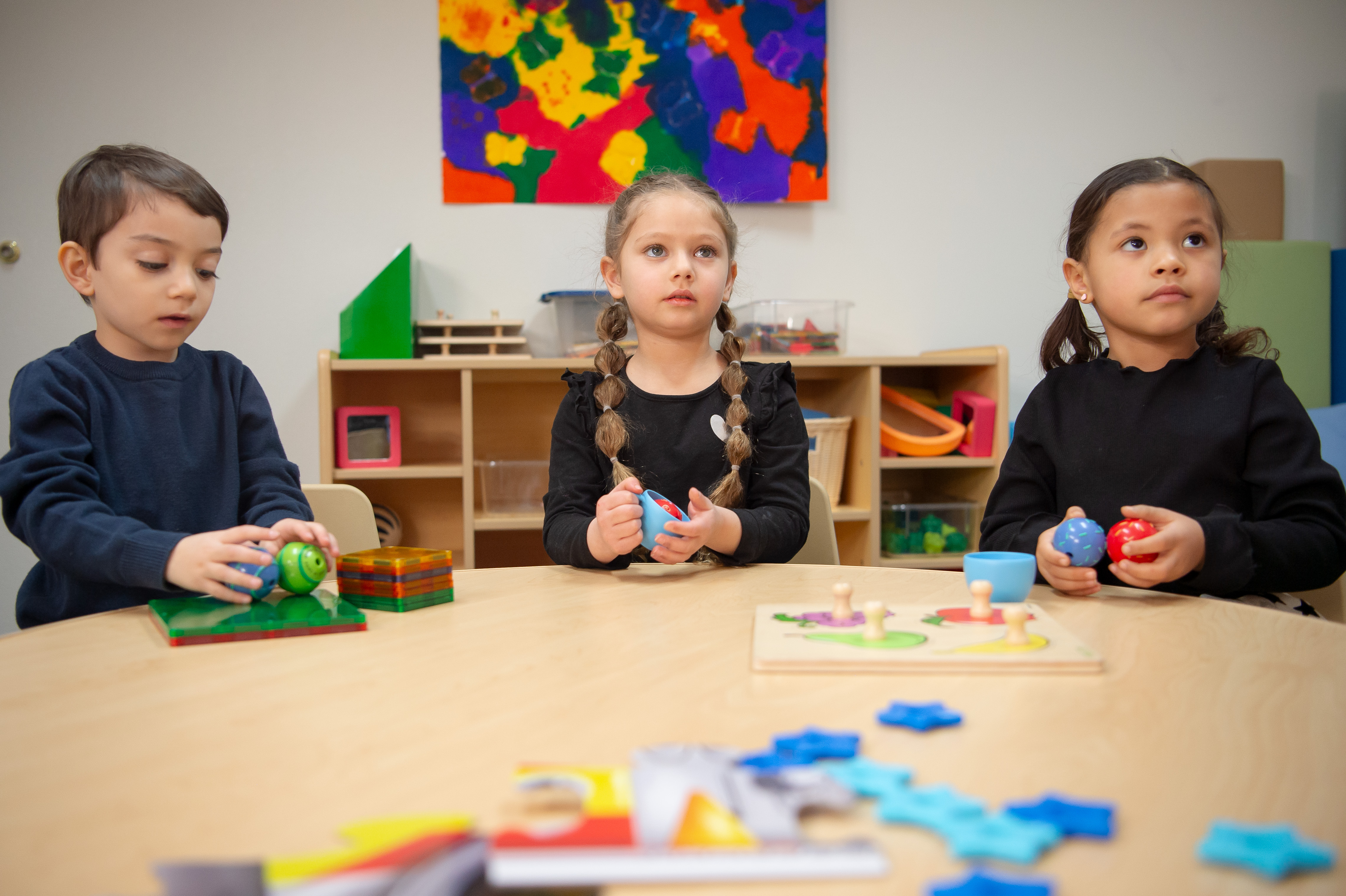 Tres niños pequeños sentados en un aula colorida alrededor de una mesa, con juguetes encima.