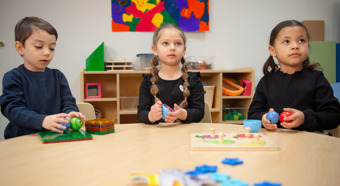 Tres niños pequeños sentados en un aula colorida alrededor de una mesa, con juguetes encima.