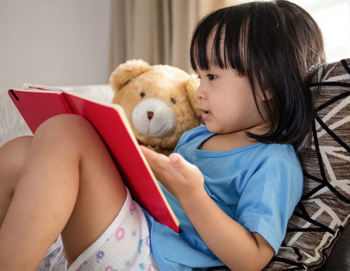 Una niña pequeña de tez marrón claro y pelo oscuro, con una remera celeste y shorts claros, sentada en un sillón leyendo un libro rojo, al lado de un oso de peluche,