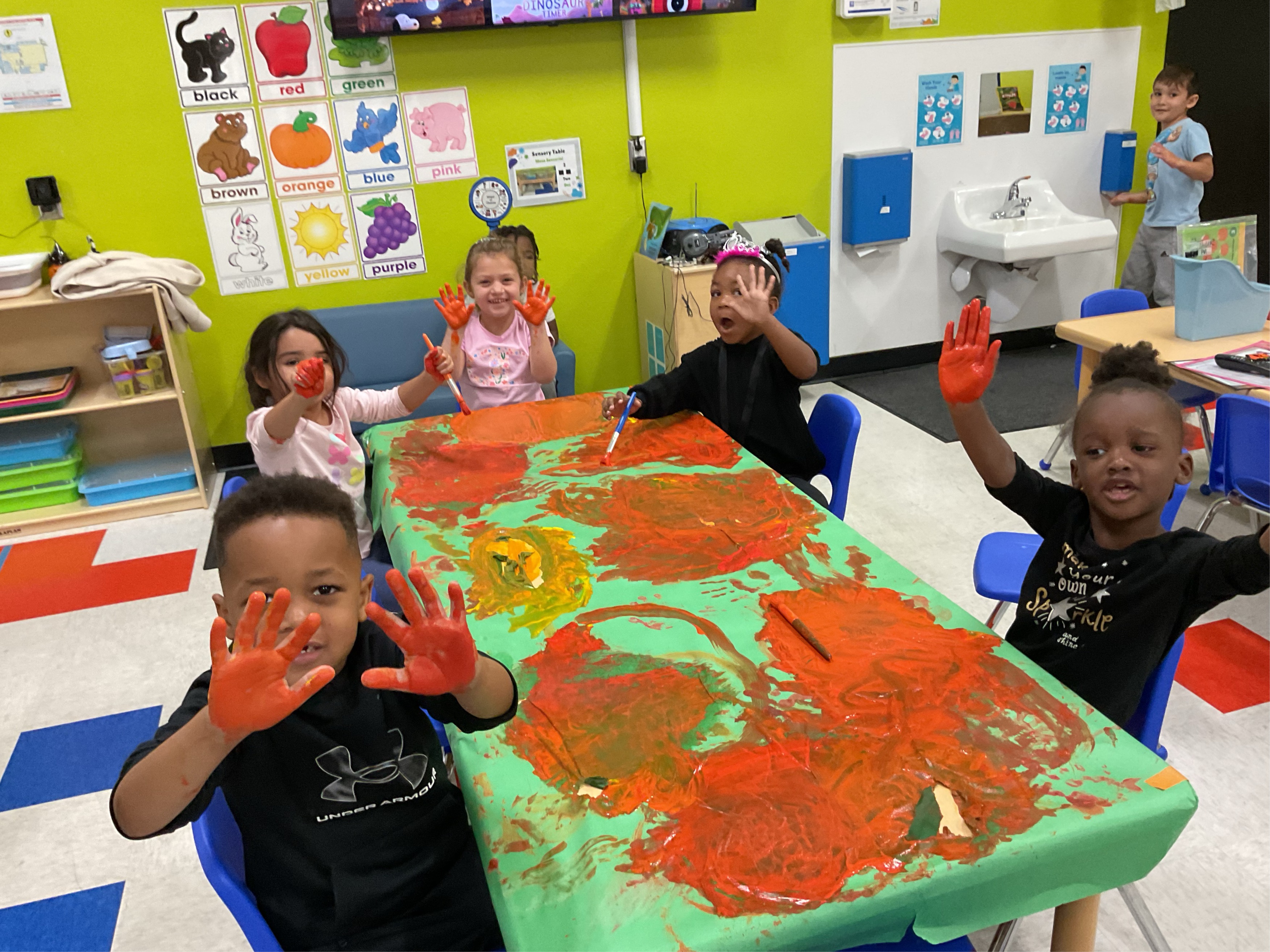 Escena en un aula con cinco niños pequeños sentados alrededor de una mesa cubierta de pintura naranja, con las manos también manchadas de pintura. Sonriendo para la foto.
