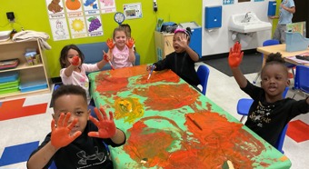Escena en un aula con cinco niños pequeños sentados alrededor de una mesa cubierta de pintura naranja, con las manos también manchadas de pintura. Sonriendo para la foto.