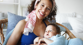 Mujer adulta sonriendo a la cámara, sosteniendo a un niño pequeño en su regazo mientras está sentada en un acogedor entorno hogareño.