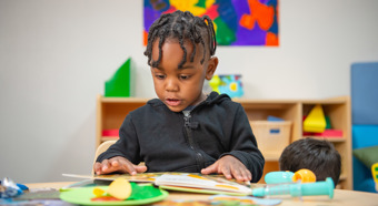 Un niño pequeño en un aula colorida mira hacia abajo y lee un libro infantil mientras está sentado.