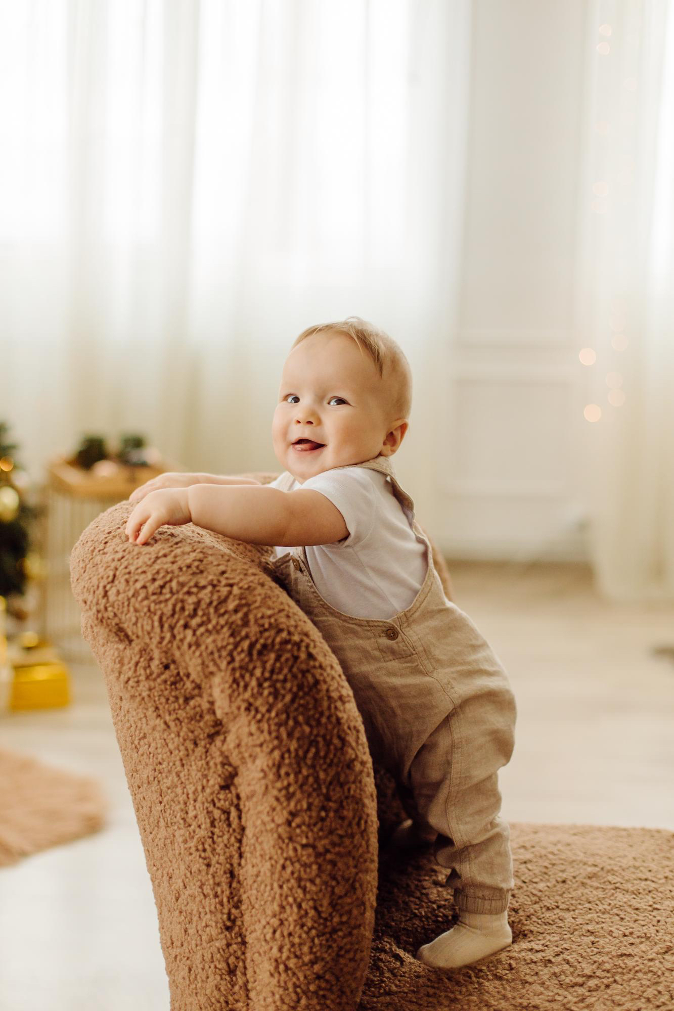 Un niño pequeño sonríe de pie sobre una silla marrón, de aspecto acogedor y peluda, en el entorno de una casa.
