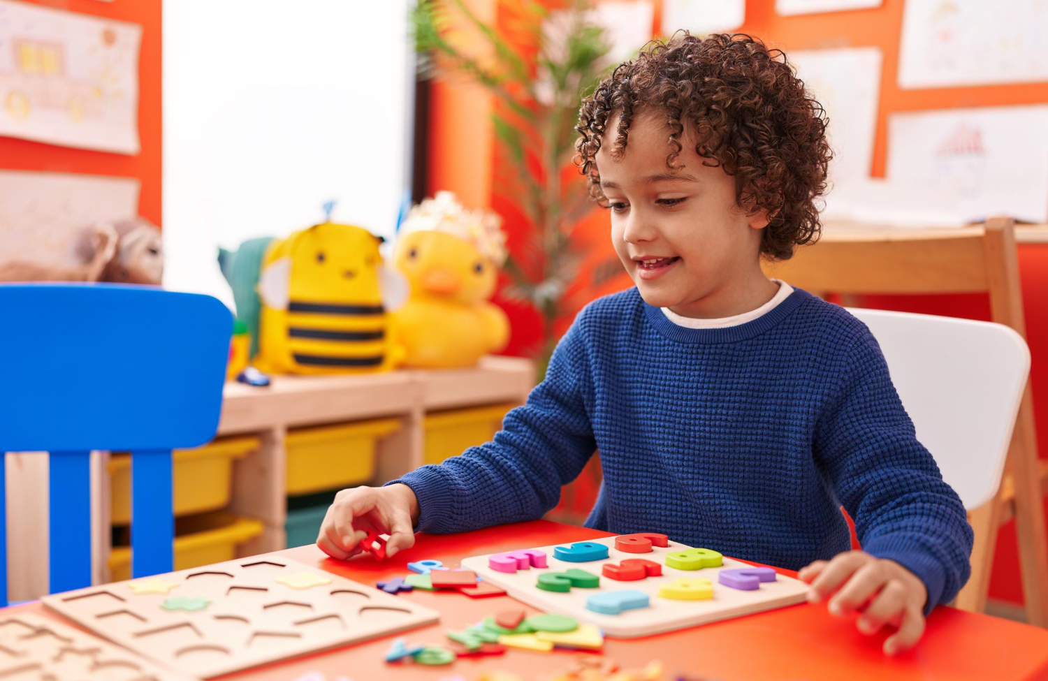 Un niño pequeño, sentado en un aula colorida, vestido con una camisa azul de manga larga, sonríe mientras juega con bloques.