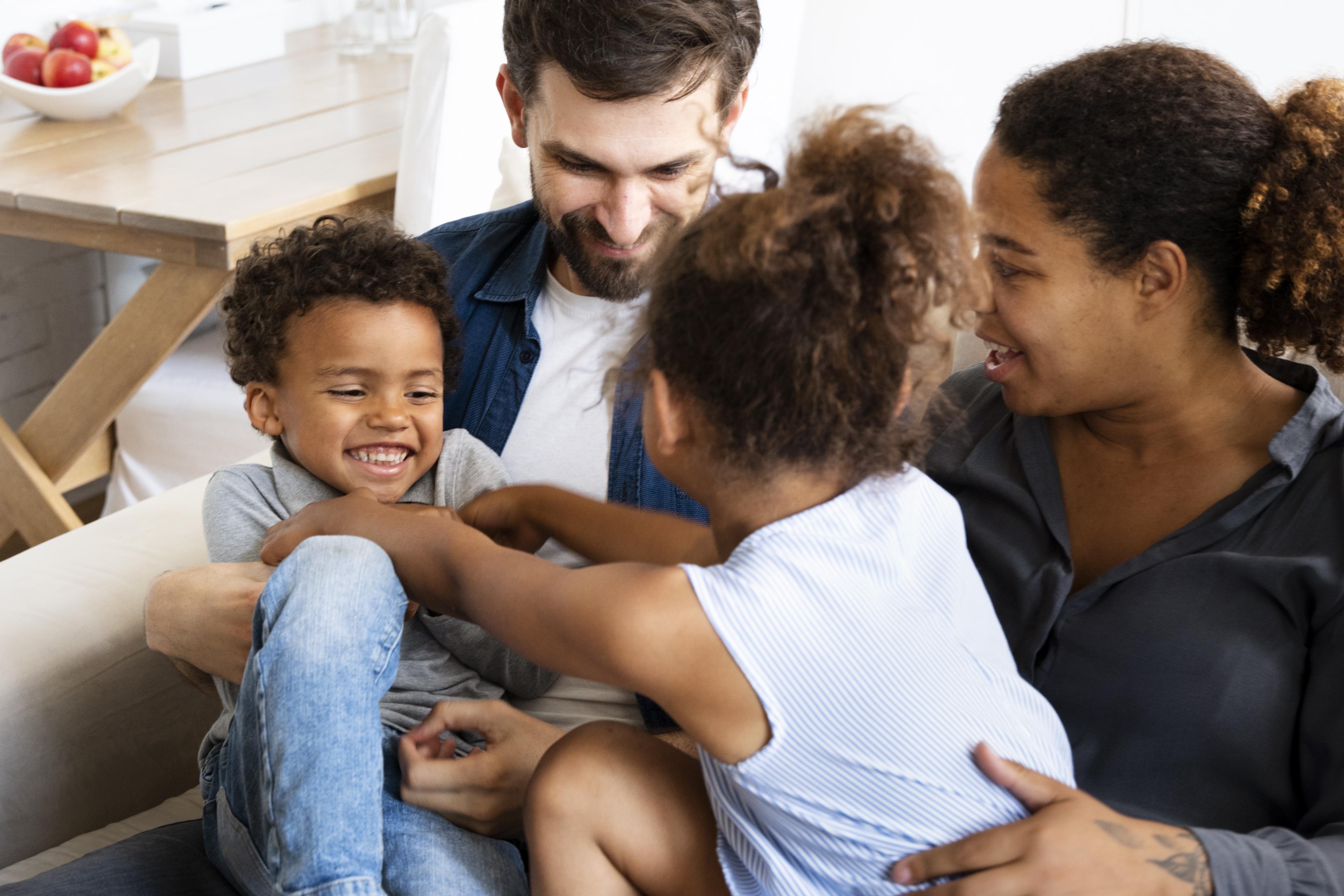 Grupo de personas, aparente una familia, conformada por dos adultos en un sillón, una mujer de tez morena y un varón de tez clara, y dos niños sentados en sus regazos, una niña de tez morena y pelo oscuro enrulado, haciéndole cosquillas a un niño más pequeño, de características similares.