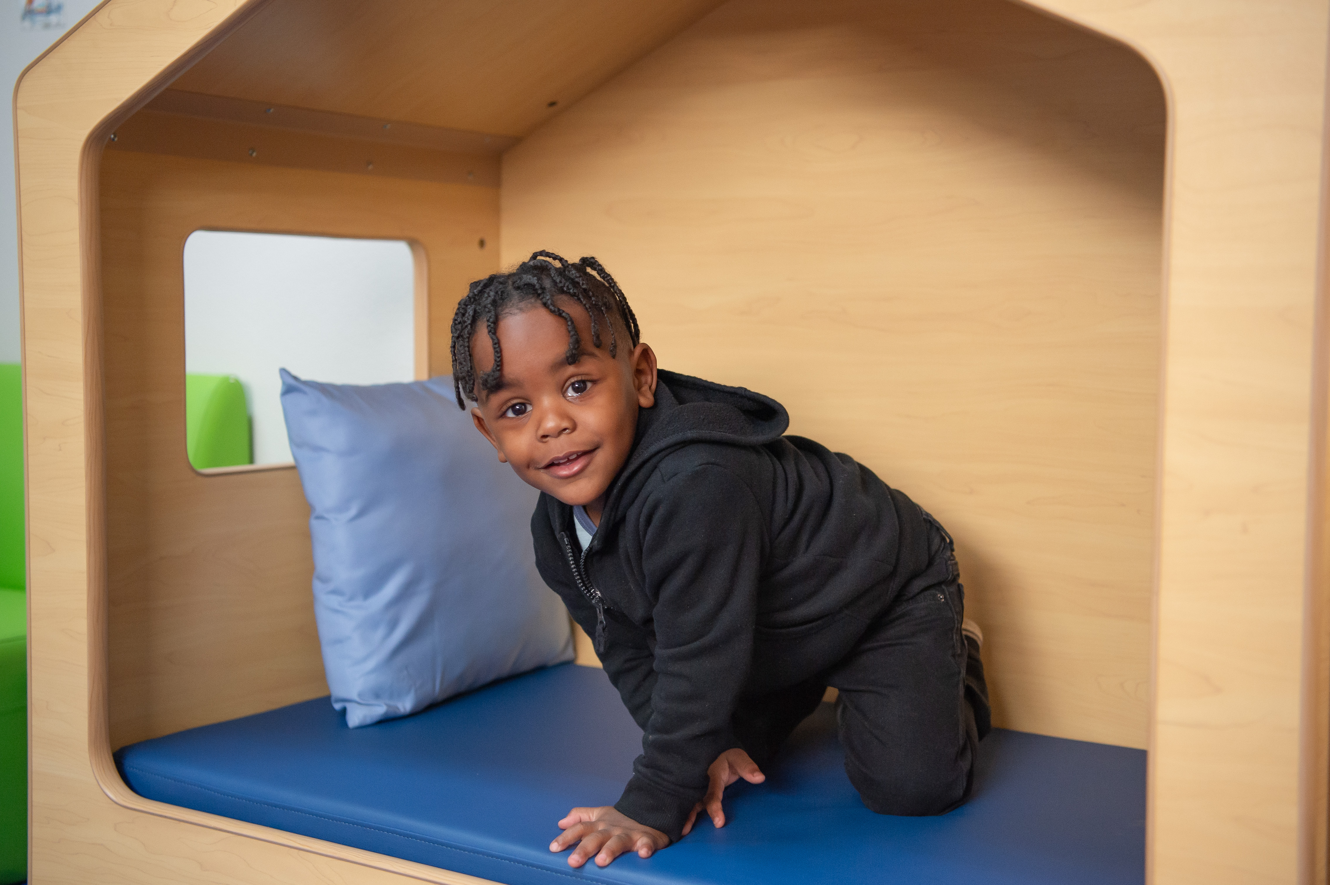 Un niño, en una casita de juegos interior con cojines azules, sonríe a la cámara. Viste ropa completamente negra.