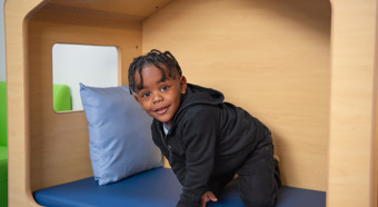 Un niño, en una casita de juegos interior con cojines azules, sonríe a la cámara. Viste ropa completamente negra.