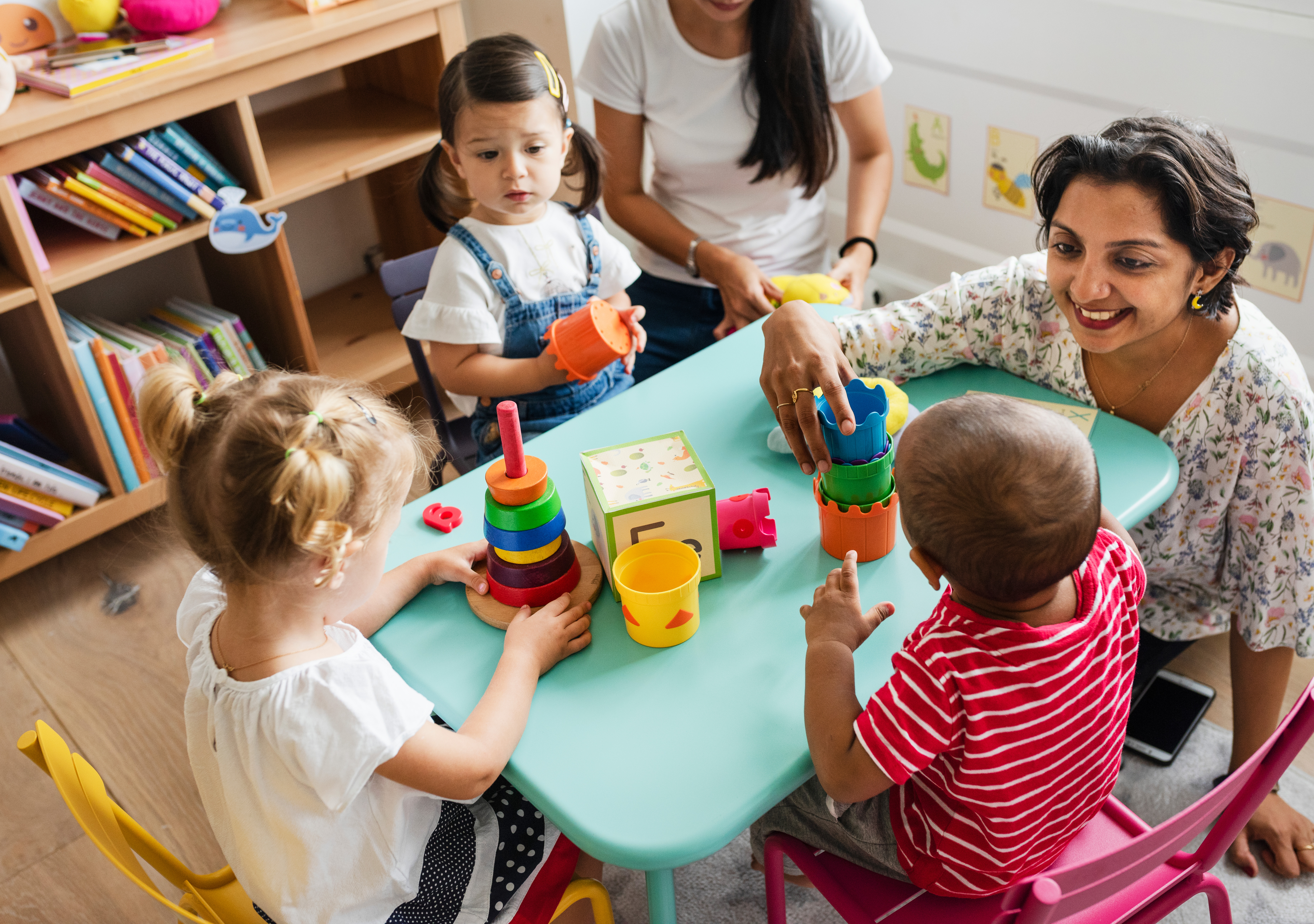 Tres niños pequeños jugando con juguetes con su maestra en una mesa de tamaño infantil.