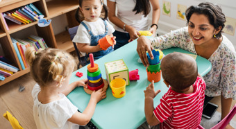 Tres niños pequeños jugando con juguetes con su maestra en una mesa de tamaño infantil.