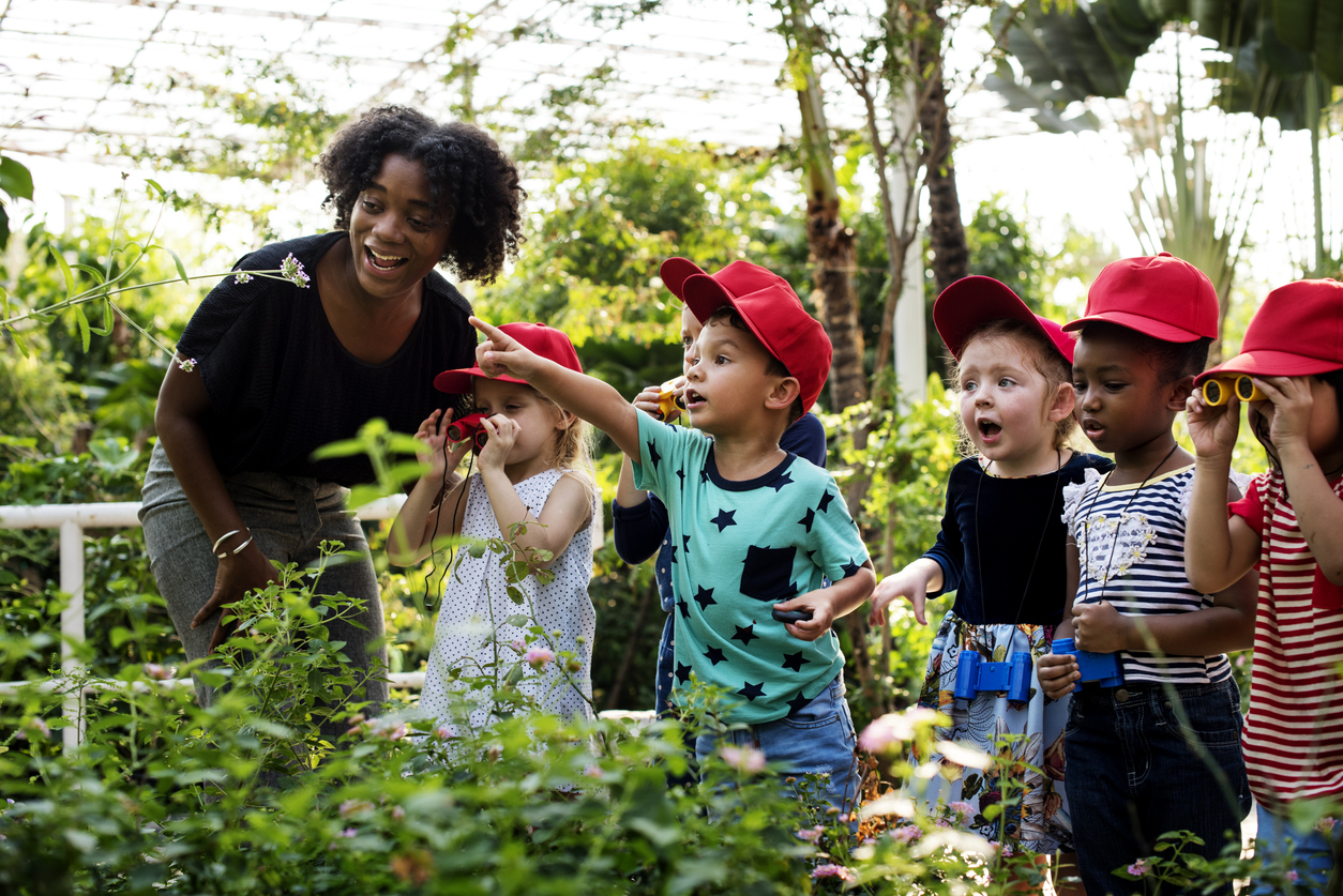 Una mujer adulta de tez y pelo oscuros, junto a un grupo de niños diverso con gorras rojas, en un vivero, mirando con sorpresa a las plantas.