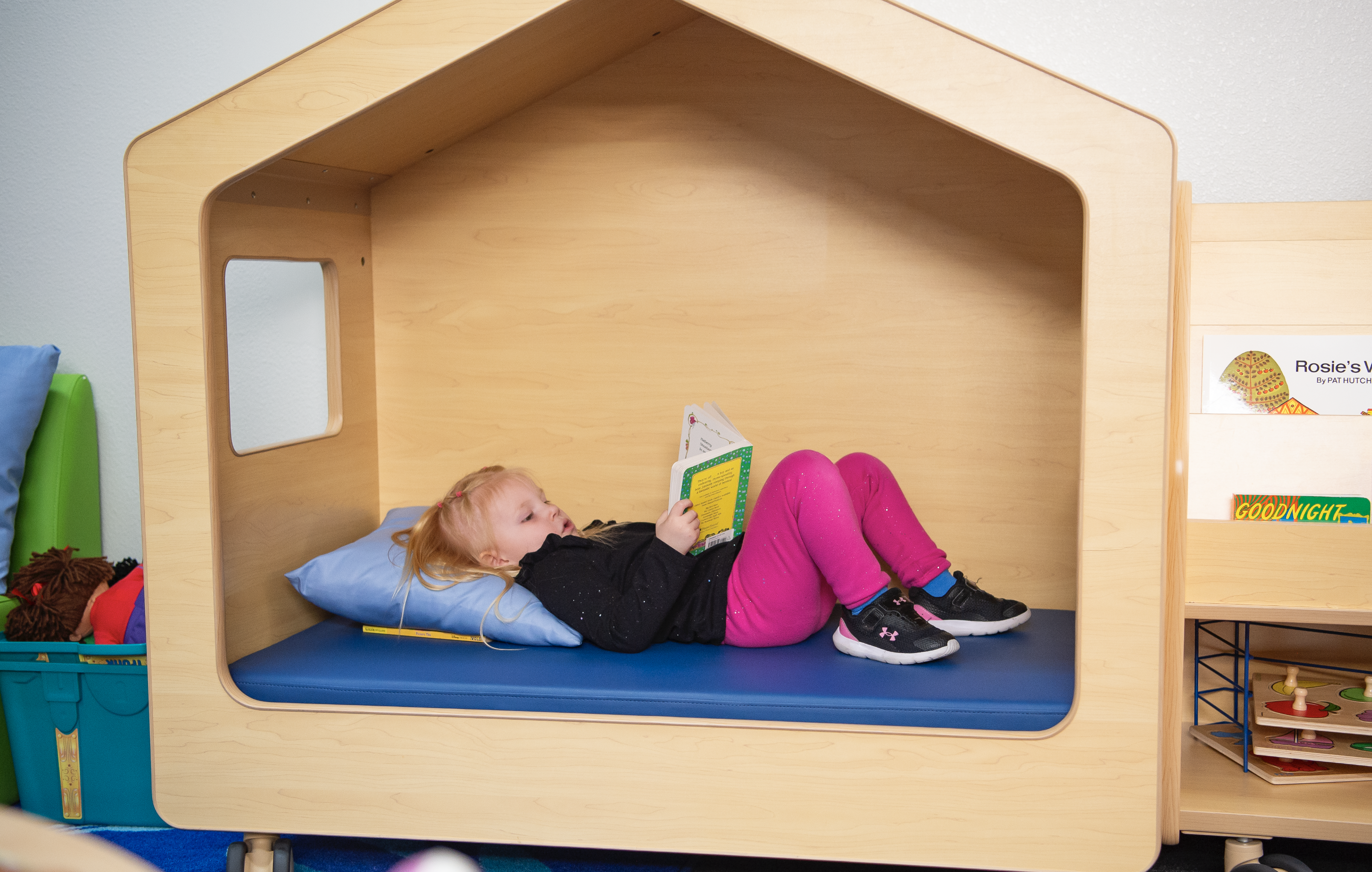 Una niña pequeña acostada en una cama con forma de casita, leyendo un libro. Lleva pantalones rosas.