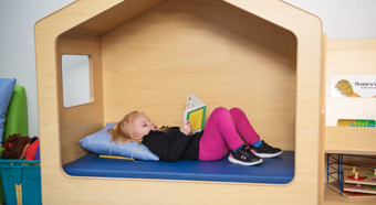 Una niña pequeña acostada en una cama con forma de casita, leyendo un libro. Lleva pantalones rosas.