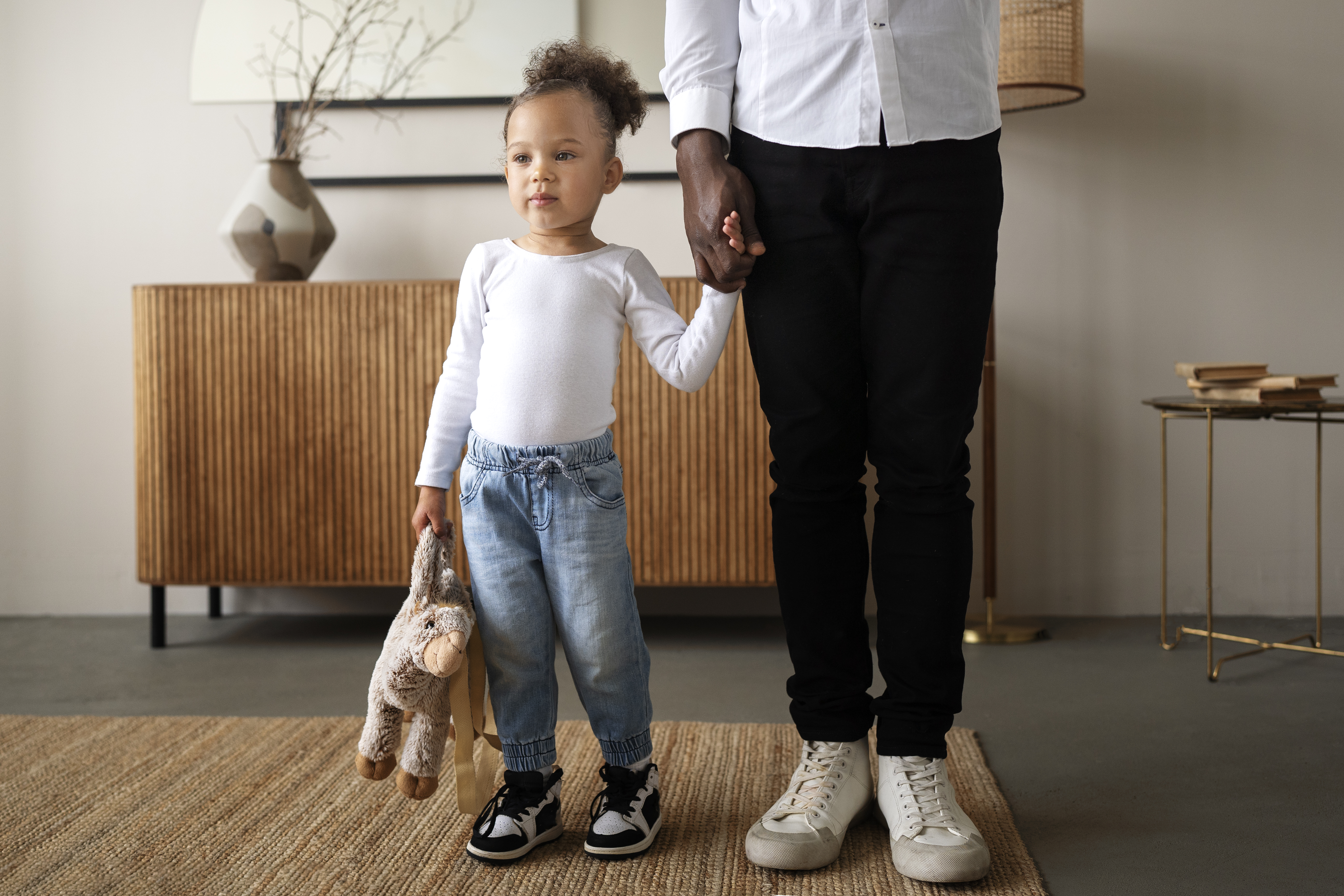 Niña con camisa blanca de manga larga y vaqueros, sosteniendo un peluche de la mano de un adulto. Interior de una casa.