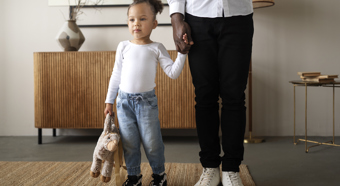 Niña con camisa blanca de manga larga y vaqueros, sosteniendo un peluche de la mano de un adulto. Interior de una casa.