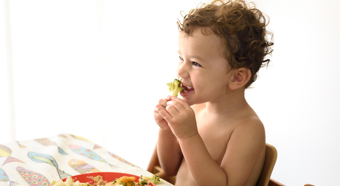 Niño pequeño sentado a la mesa, comiendo un trozo de brócoli, sonriendo y riendo.