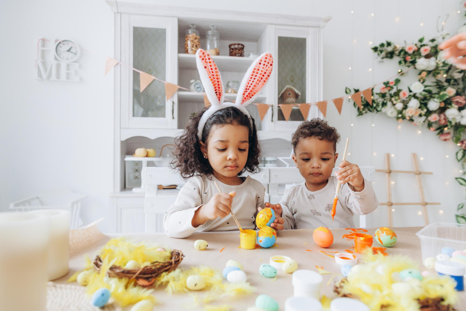 Dos niños pequeños sentados a una mesa en un entorno hogareño, pintando huevos de Pascua; uno de ellos lleva puestas unas orejas de conejo de Pascua.