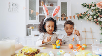 Dos niños pequeños sentados a una mesa en un entorno hogareño, pintando huevos de Pascua; uno de ellos lleva puestas unas orejas de conejo de Pascua.