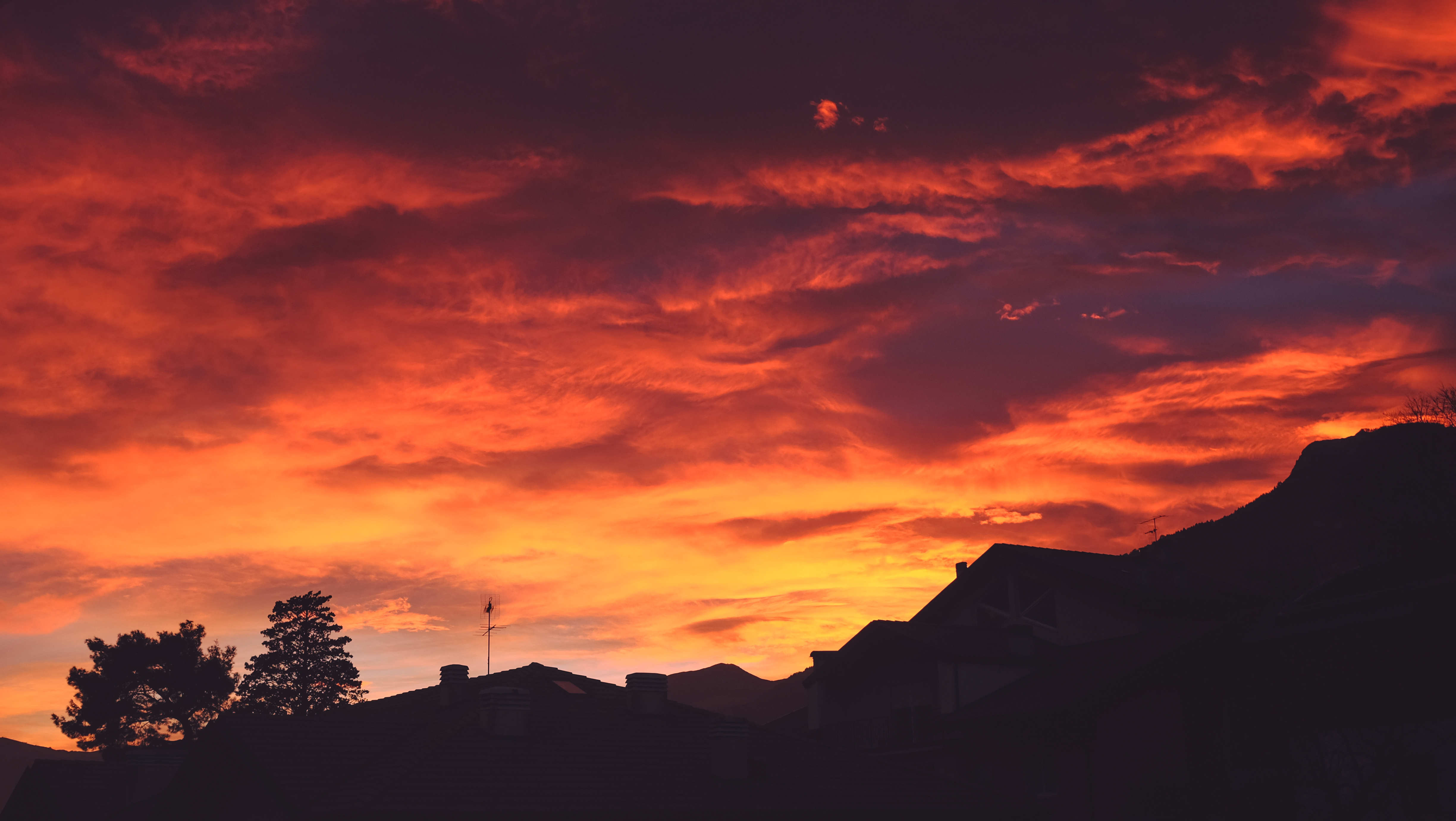 Cielo de atardecer con tonos rojos, naranjas y amarillos sobre un barrio con montañas y árboles en primer plano y fondo.