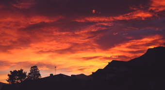 Cielo de atardecer con tonos rojos, naranjas y amarillos sobre un barrio con montañas y árboles en primer plano y fondo.