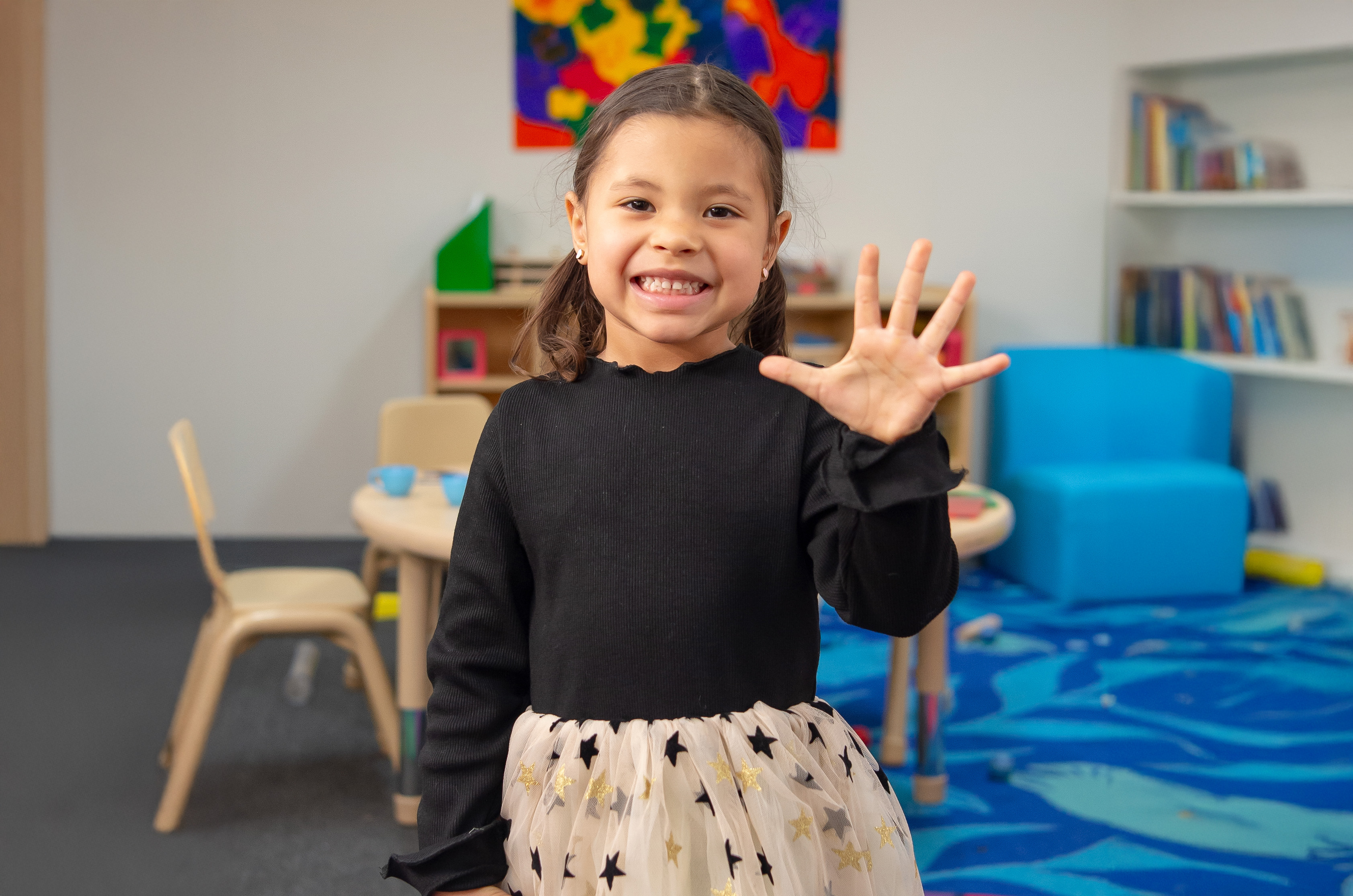 Una niña sonríe a la cámara y levanta la mano con cinco dedos, en un aula colorida