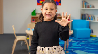 Una niña sonríe a la cámara y levanta la mano con cinco dedos, en un aula colorida