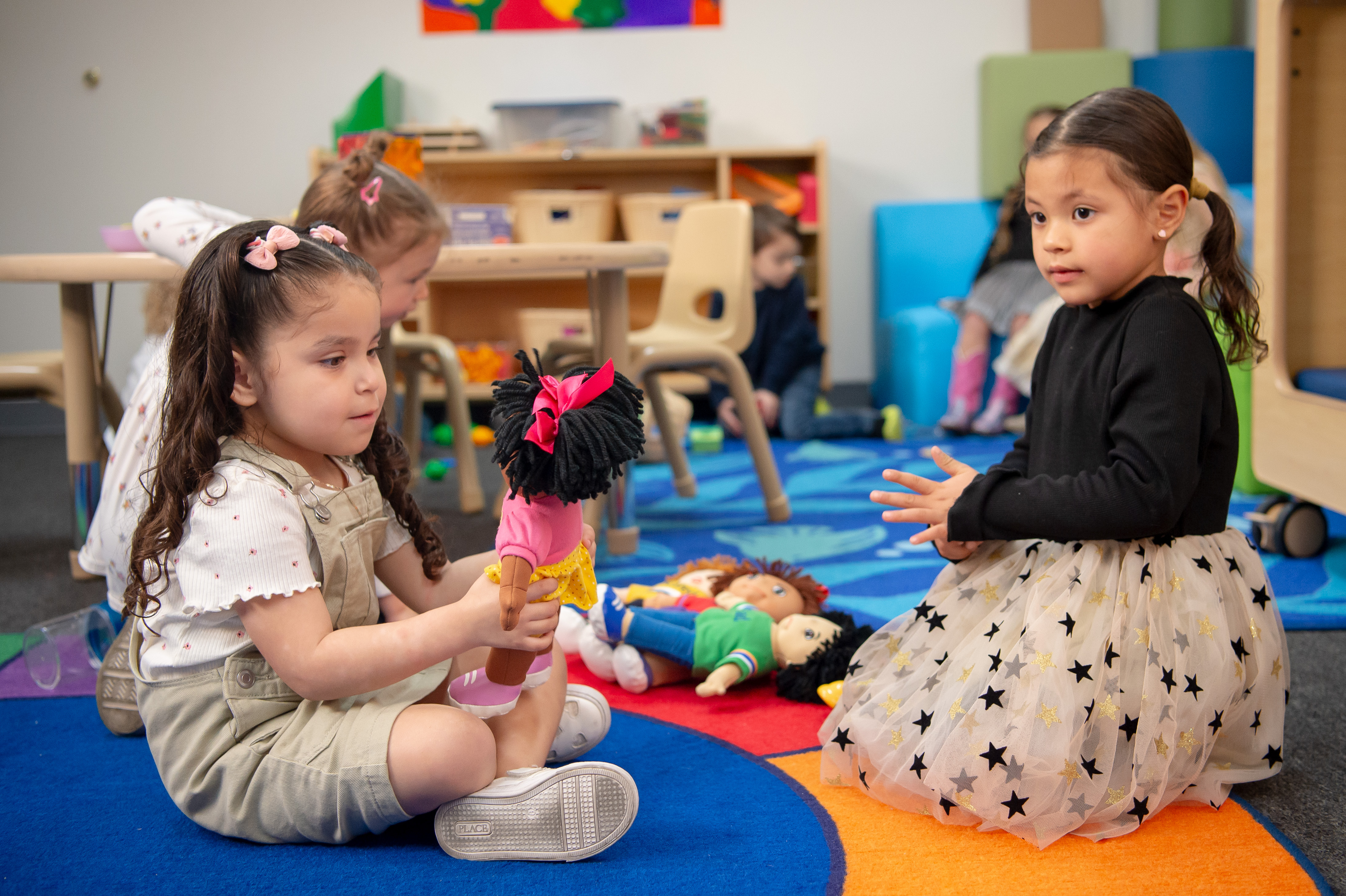 Dos niñas hispanas sentadas en un colorido suelo alfombrado y jugando con muñecas.