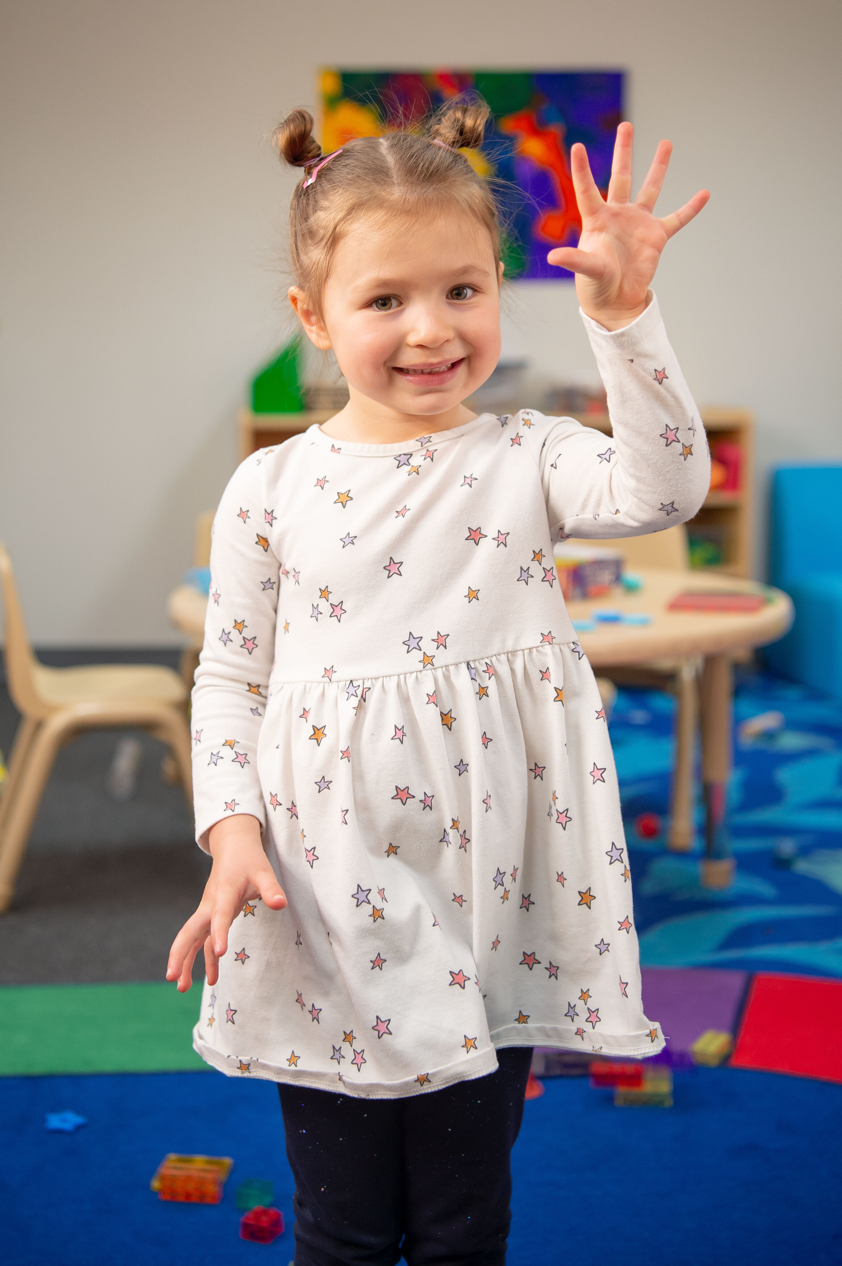 Una niña pequeña de tez clara y rubia con el pelo atado, con un vestido claro, sonriéndo y mostrándole los cinco dedos de su mano extendida a la cámara.